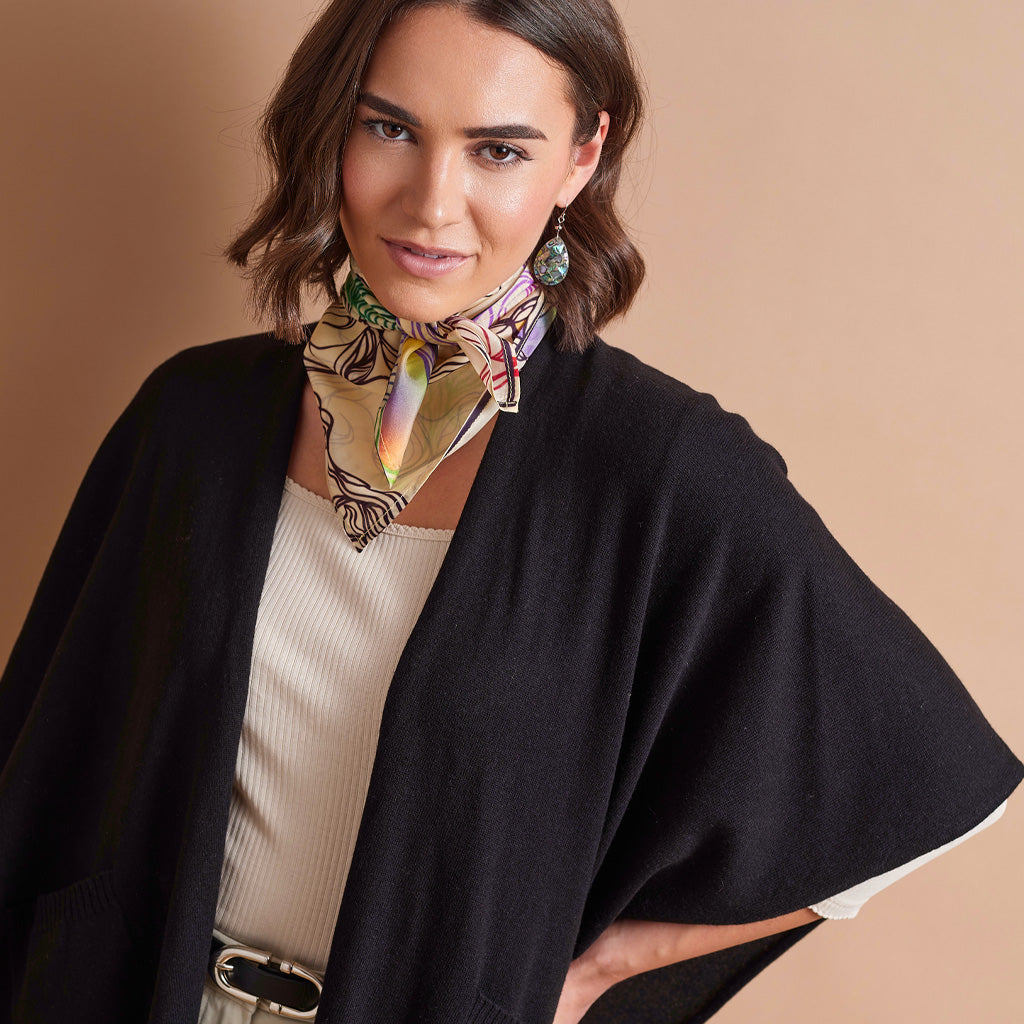 A woman wearing a black open-front cardigan over a cream ribbed top, accessorized with a multicolor patterned silk neck scarf and green statement earrings, set against a solid beige background.