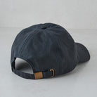 A dark navy blue cotton baseball cap shown from the back, featuring a matching fabric strap with a gold-tone metal buckle and a small tan leather accent.