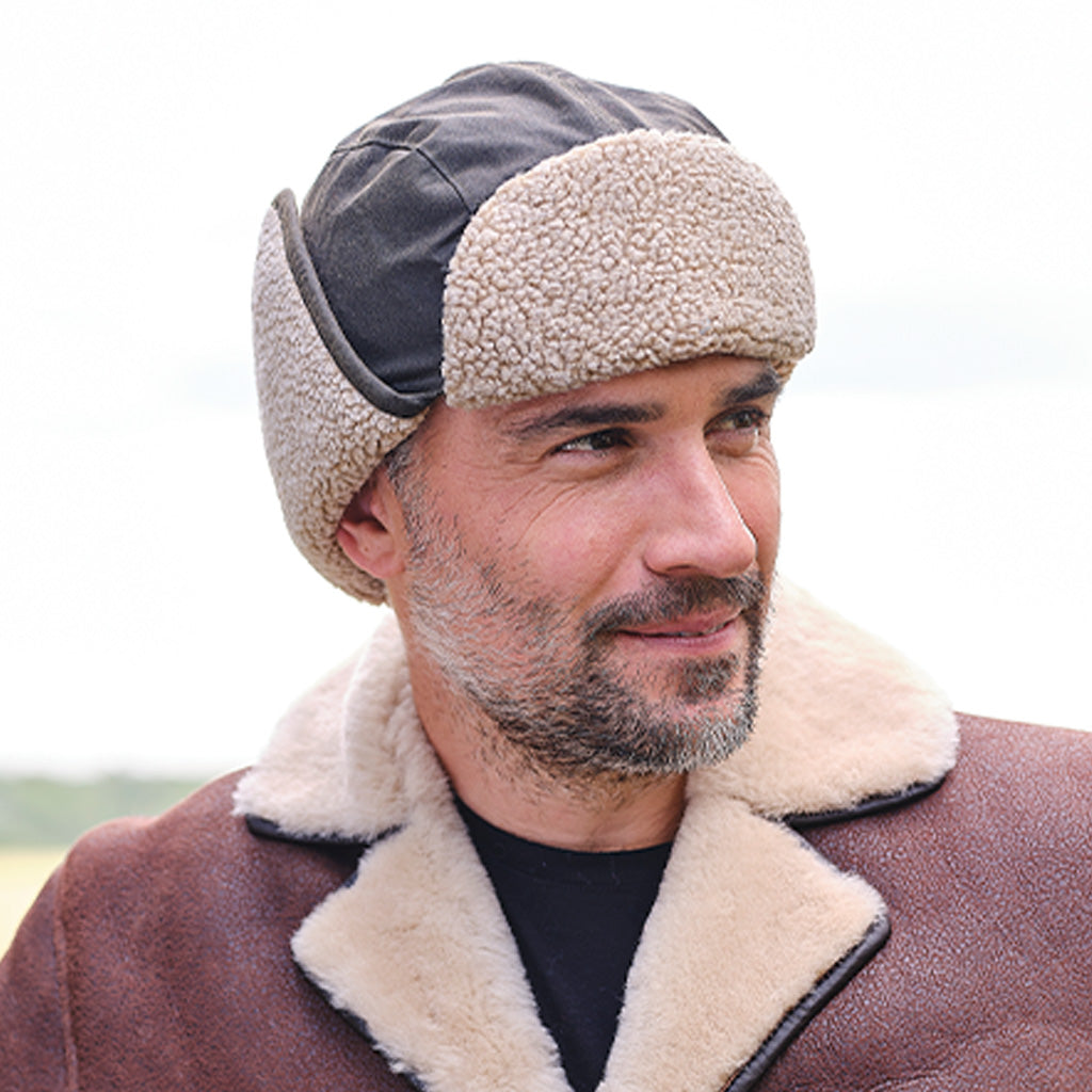 A man wearing a brown shearling-lined trapper hat with a dark gray top panel and a brown shearling-lined collar with black piping over a black shirt, photographed outdoors against a bright, overexposed sky.