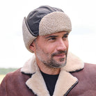 A man wearing a brown shearling-lined trapper hat with a dark gray top panel and a brown shearling-lined collar with black piping over a black shirt, photographed outdoors against a bright, overexposed sky.