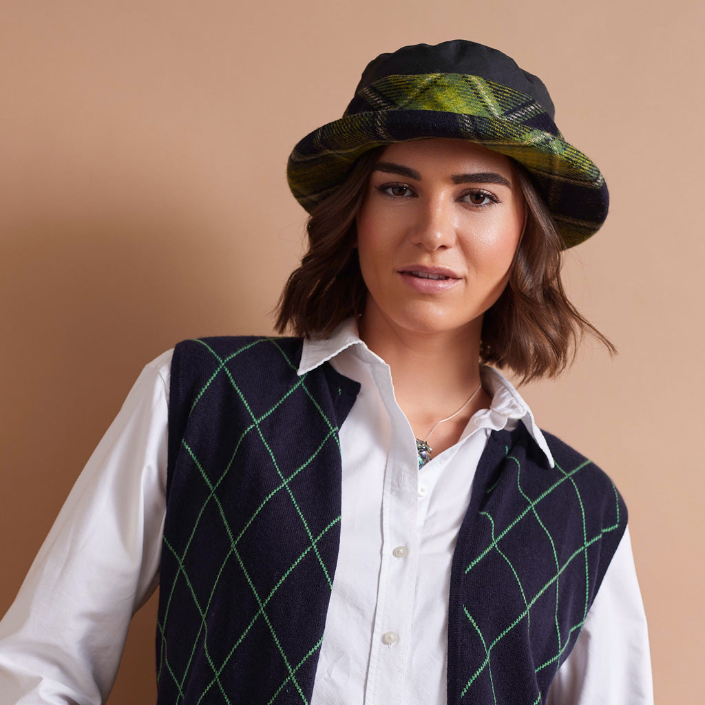 A woman wearing a dark navy vest with a green diamond argyle pattern, a white collared shirt, and a black bucket hat with a green and yellow plaid brim, set against a solid beige background.