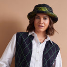 A woman wearing a dark navy vest with a green diamond argyle pattern, a white collared shirt, and a black bucket hat with a green and yellow plaid brim, set against a solid beige background.