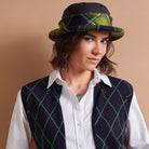 A woman wearing a black and green plaid bowler hat, a dark navy argyle vest with green lines over a white collared shirt, and a necklace, set against a solid warm beige background.