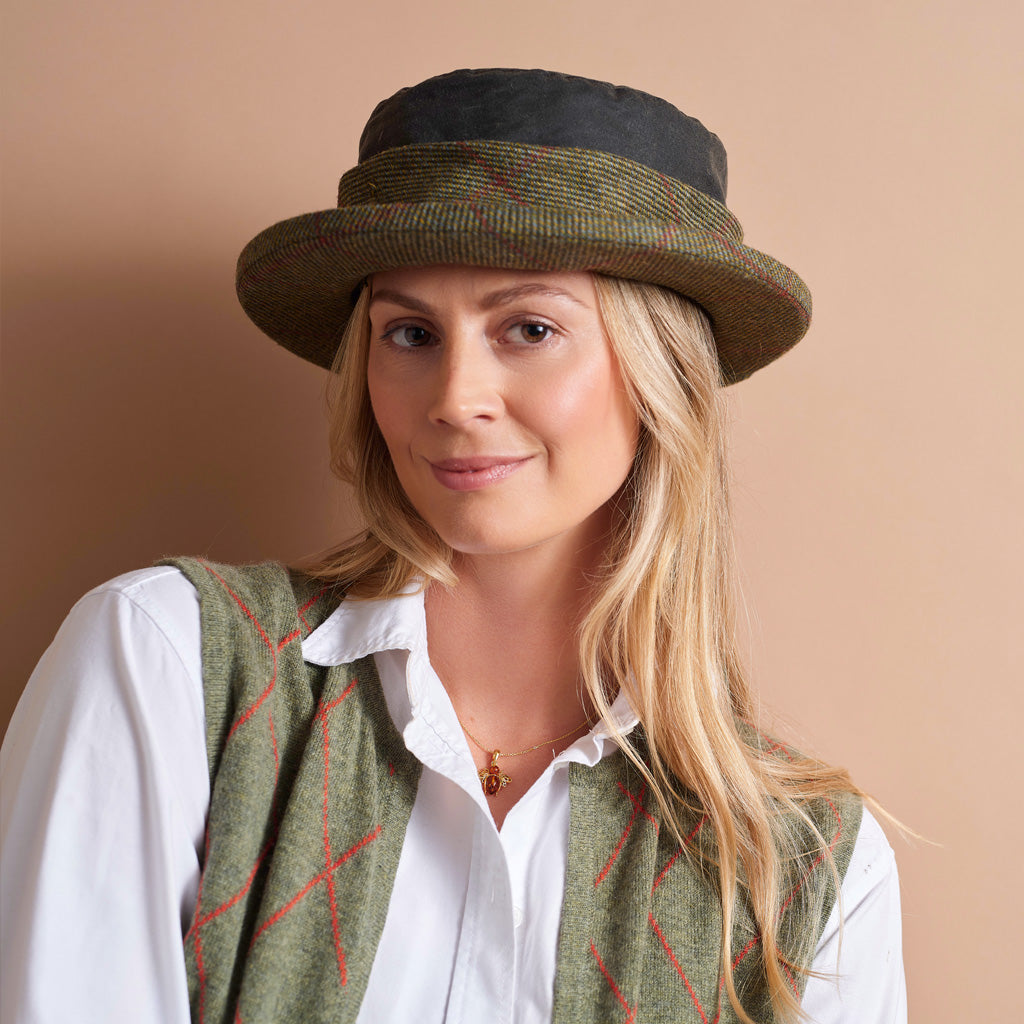 A woman wearing a textured green tweed bowler hat with a black top band, styled with a white collared shirt and a matching green tweed vest with red crisscross lines, set against a solid beige background.