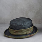 A dark charcoal gray felt hat with a textured olive green and red tweed band, displayed on a neutral gray surface against a textured gray background.