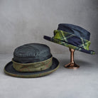 Two dark navy blue felt hats with green and yellow plaid wool bands, one displayed flat on a surface and the other standing on a metallic stand against a gray background.