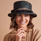 A smiling woman wearing a black wide-brim hat with a textured brown band and a camel-colored knit top, set against a solid beige background.