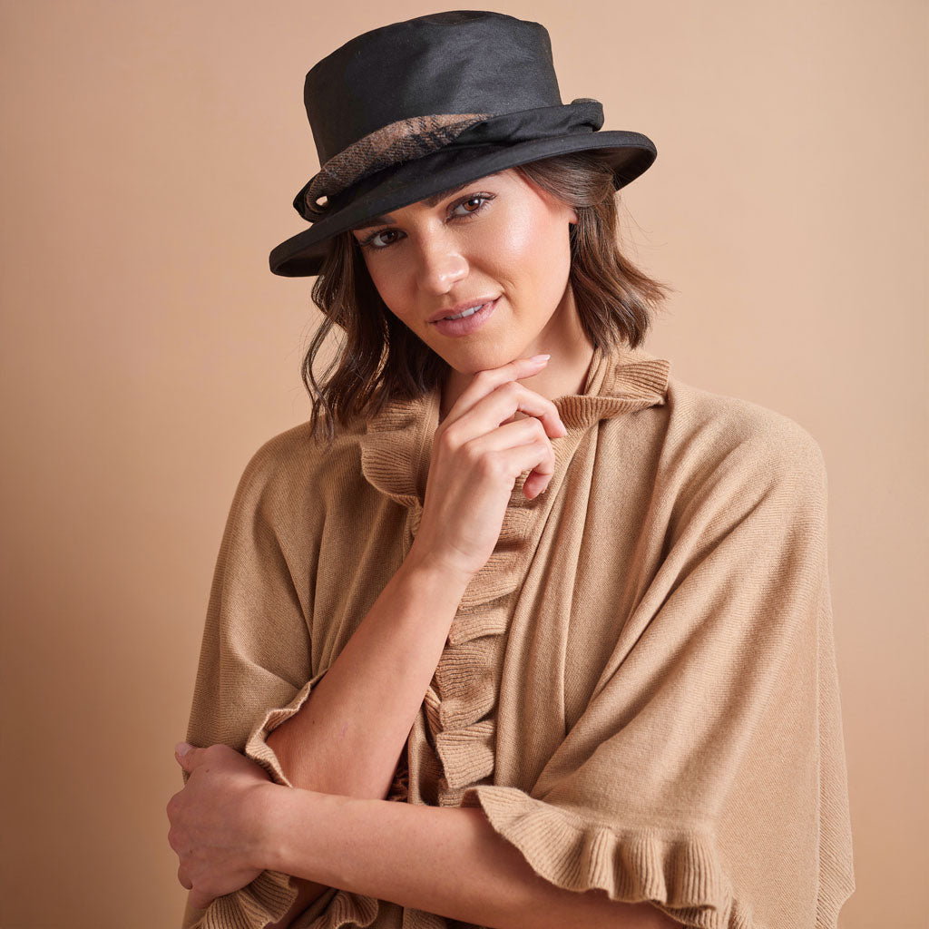 A woman wearing a black felt fedora hat with a patterned band and a beige ruffled knit poncho, posed against a solid beige studio background.