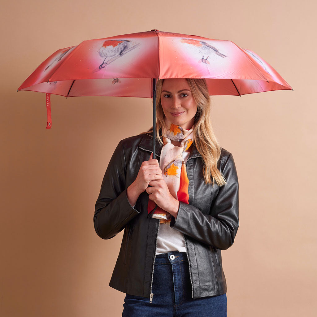 A woman holding a red umbrella with a bird pattern, wearing a black leather jacket, white shirt, and orange scarf, against a solid beige background.