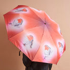 A pink and orange umbrella with printed red robin bird illustrations, held open by a person against a plain beige background.