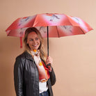 A woman holding a pink umbrella with a bird pattern, wearing a black leather jacket and a white scarf with orange and pink details, set against a solid beige background.